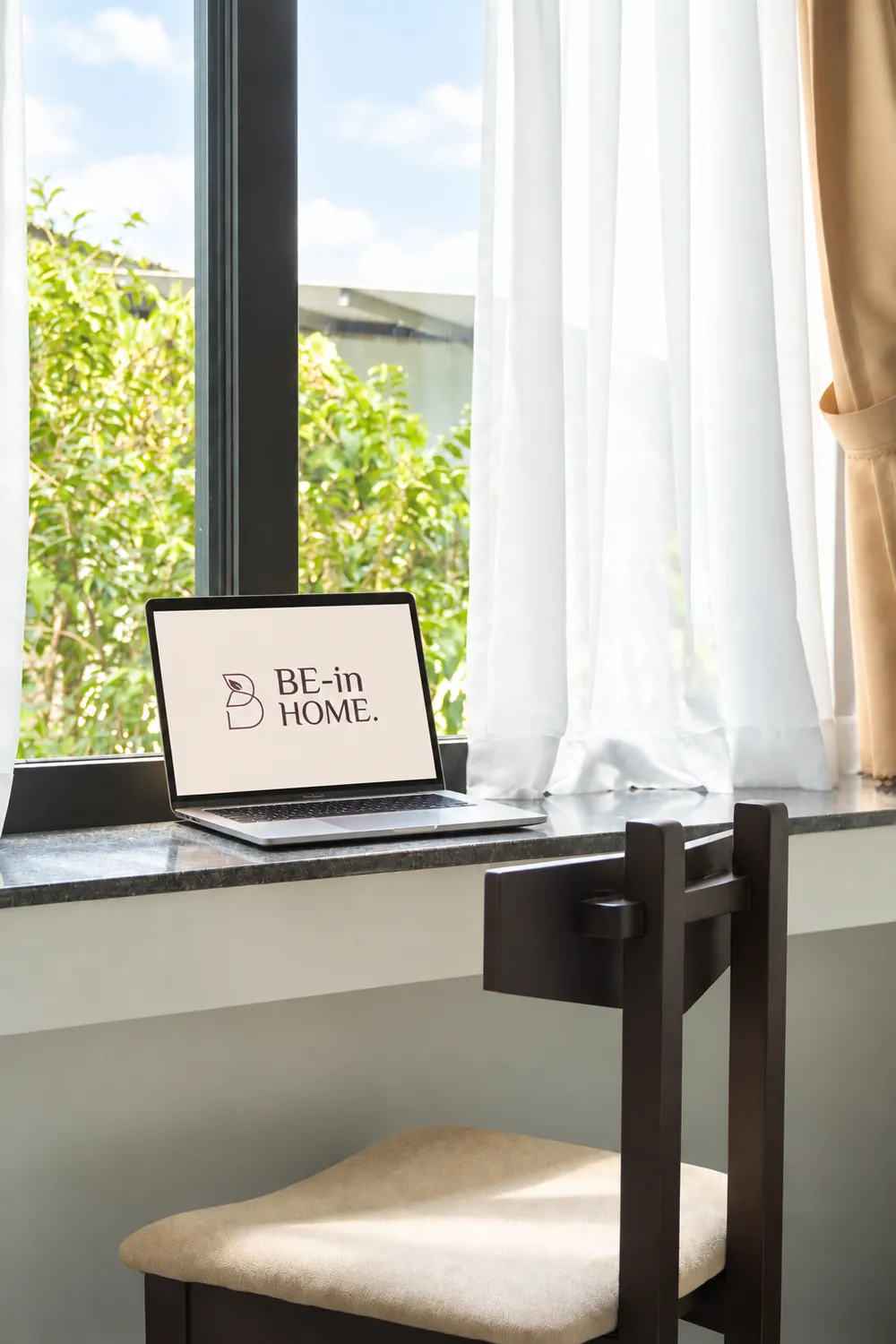 A MacBook displaying the Be-in Home logo at the sunlit window workspace.